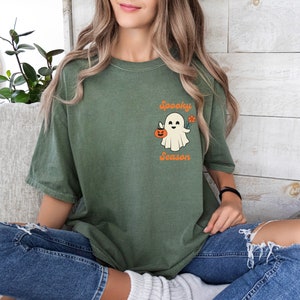 May include: A green t-shirt with a white ghost holding a jack-o-lantern and a flower. The text "Spooky Season" is printed below the ghost.