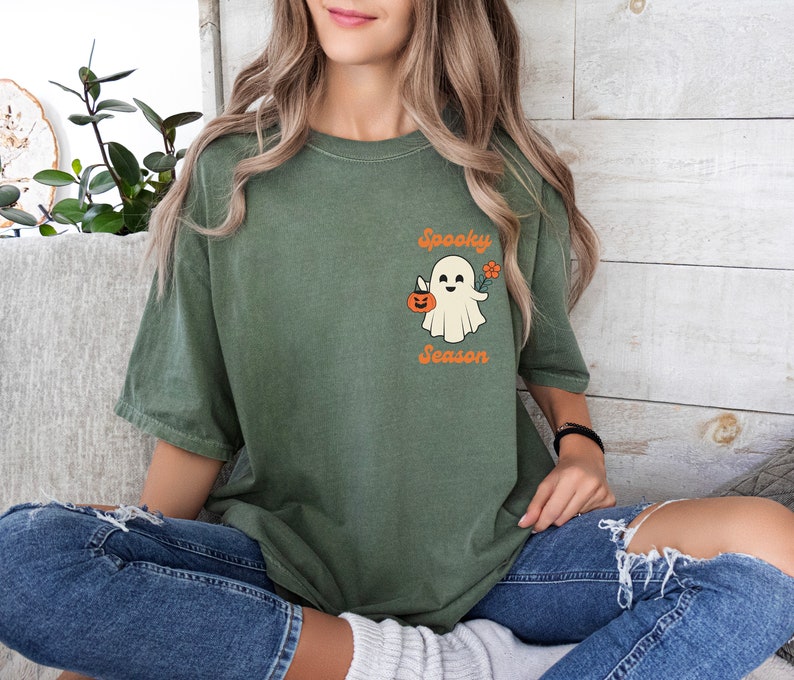 May include: A green t-shirt with a white ghost holding a jack-o-lantern and a flower. The text "Spooky Season" is printed below the ghost.