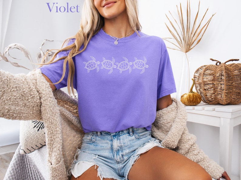 May include: A purple t-shirt with a row of five white sea turtles printed on the front. The shirt is worn by a person with long blonde hair and is styled with denim shorts.
