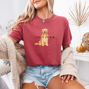 May include: A woman wearing a maroon t-shirt with a golden retriever illustration and the text "Life is Golden". She is wearing denim shorts and a white fuzzy cardigan.
