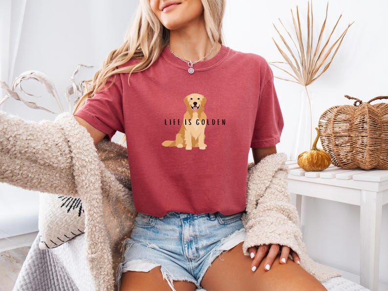 May include: A woman wearing a maroon t-shirt with a golden retriever illustration and the text "Life is Golden". She is wearing denim shorts and a white fuzzy cardigan.