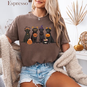 May include: Espresso-colored t-shirt featuring a Halloween-themed graphic. The design showcases four black Labrador dogs, each adorned with festive accessories like witch hats and pumpkin-shaped treat containers. The text "Trick or Treat" is also visible.