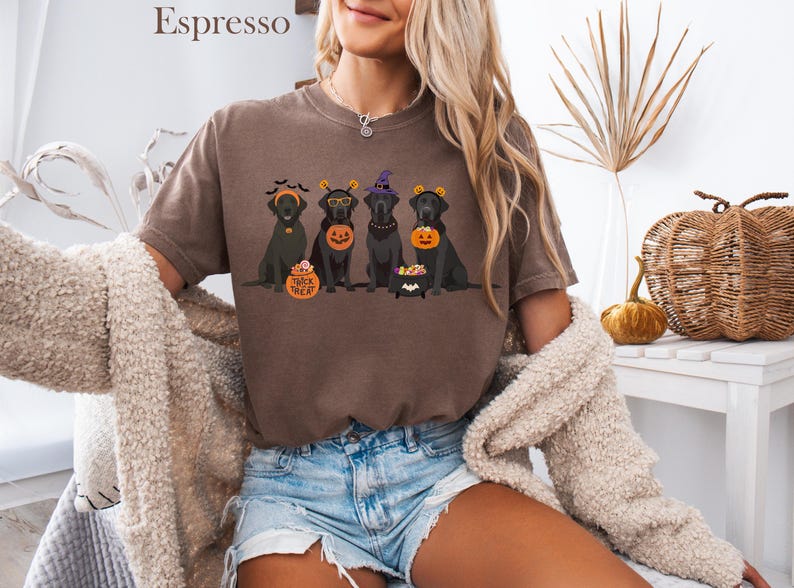 May include: Espresso-colored t-shirt featuring a Halloween-themed graphic. The design showcases four black Labrador dogs, each adorned with festive accessories like witch hats and pumpkin-shaped treat containers. The text "Trick or Treat" is also visible.