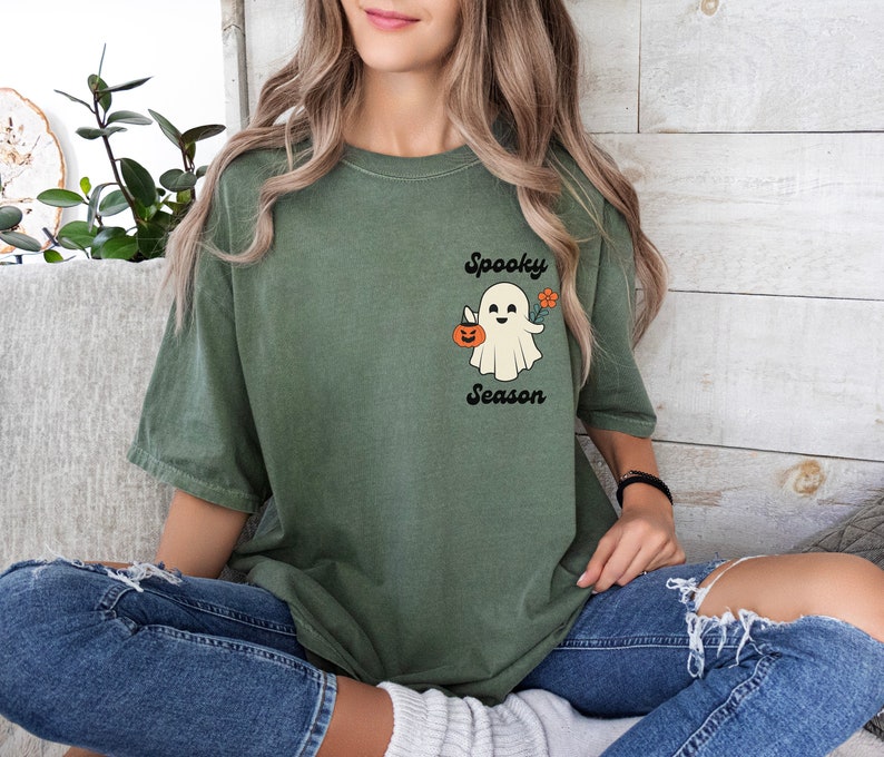 May include: A green t-shirt with a white ghost holding a pumpkin and a flower. The text "Spooky Season" is printed below the ghost.