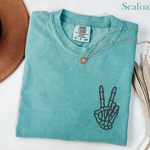Puede incluir: Camiseta verde mar con un gráfico de la mano de un esqueleto haciendo el signo de la paz. La camiseta tiene cuello redondo y mangas cortas. Un collar dorado con un colgante está drapeado sobre la camiseta. La camiseta está doblada sobre una superficie blanca.
