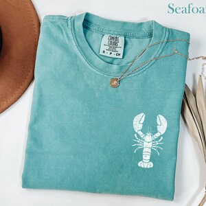 以下が含まれることがあります： 白のロブスターのグラフィックが描かれたシーフォームグリーンのTシャツ。Tシャツはクルーネックと半袖です。ブランド名「COMFORT COLORS」はタグに表示されています。