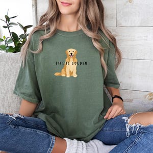 May include: A woman wearing a green t-shirt with a golden retriever illustration and the text "Life is Golden". She is wearing blue jeans and sitting on a couch.