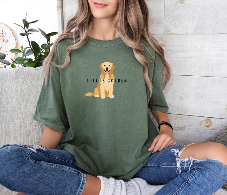 May include: A woman wearing a green t-shirt with a golden retriever illustration and the text "Life is Golden". She is wearing blue jeans and sitting on a couch.
