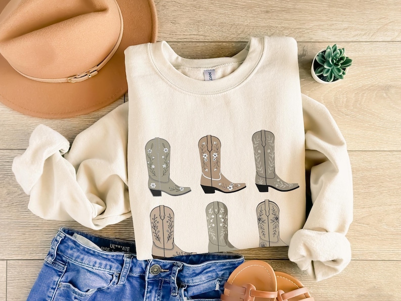 May include: A cream colored sweatshirt with a graphic of six brown cowboy boots with floral embroidery. The boots are arranged in two rows of three.