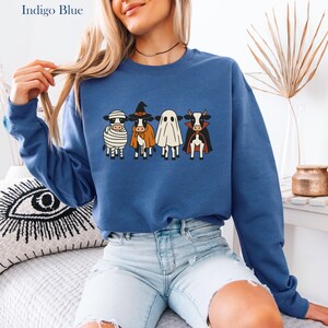 May include: Indigo blue sweatshirt featuring a Halloween-themed design. The design includes four cartoon cows dressed as a mummy, witch, ghost, and vampire. The sweatshirt is a crew neck style and is made of a soft material.