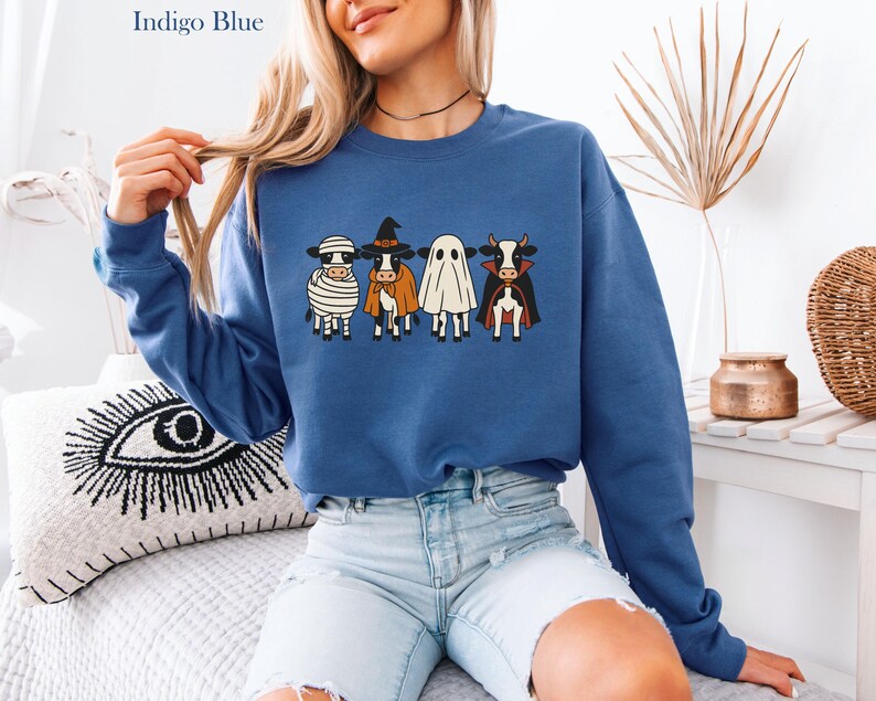 May include: Indigo blue sweatshirt featuring a Halloween-themed design. The design includes four cartoon cows dressed as a mummy, witch, ghost, and vampire. The sweatshirt is a crew neck style and is made of a soft material.