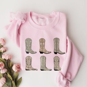 May include: Pink sweatshirt with a graphic of six brown cowboy boots with floral embroidery.
