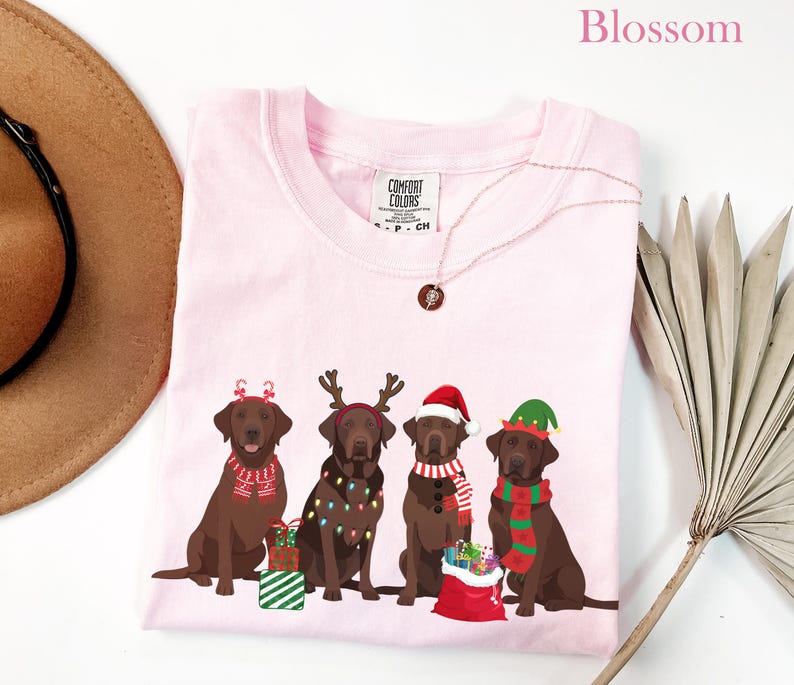 Op de afbeelding: Een lichtroze T-shirt met een afbeelding van vier bruine Labrador Retrievers met kerstaccessoires. De honden dragen kerstmutsen, rendiergeweien en elfenmutsen. Het woord "Blossom" staat bovenaan.