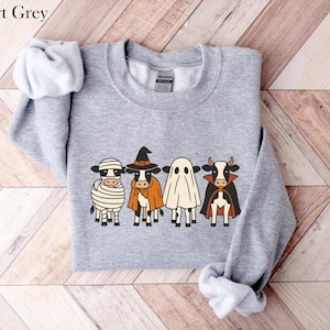 May include: Sport Grey sweatshirt featuring a Halloween-themed design. The graphic depicts four cartoon cows dressed as a mummy, witch, ghost, and vampire. The sweatshirt is a light gray color.