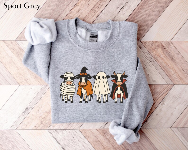 May include: Sport Grey sweatshirt featuring a Halloween-themed design. The graphic depicts four cartoon cows dressed as a mummy, witch, ghost, and vampire. The sweatshirt is a light gray color.