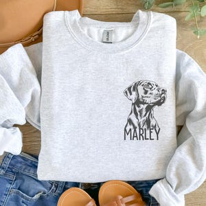 Op de afbeelding: Een grijs sweatshirt met een zwart-witte illustratie van een hondenkop en de naam "MARLEY" eronder gedrukt.