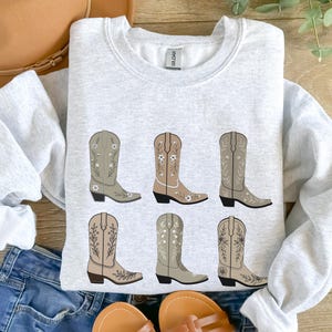 May include: A gray sweatshirt with a graphic of six brown cowboy boots with floral embroidery. The boots are arranged in two rows of three.