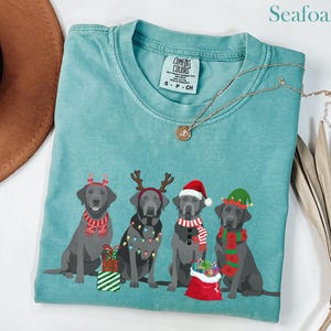 May include: Seafoam green t-shirt featuring four gray Labrador Retrievers dressed in Christmas attire. Each dog has a festive accessory, including antlers, a Santa hat, and elf ears. The shirt has a Comfort Colors tag.