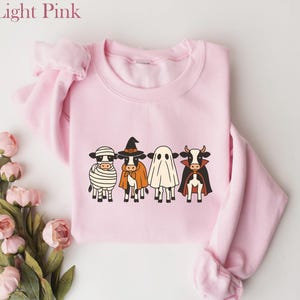 May include: Light pink sweatshirt featuring a Halloween-themed design. The graphic depicts four cows dressed as a mummy, witch, ghost, and vampire. The sweatshirt has long sleeves and a crew neck. The words "Light Pink" are in the upper left corner.