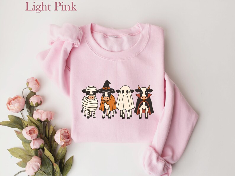 May include: Light pink sweatshirt featuring a Halloween-themed design. The graphic depicts four cows dressed as a mummy, witch, ghost, and vampire. The sweatshirt has long sleeves and a crew neck. The words "Light Pink" are in the upper left corner.