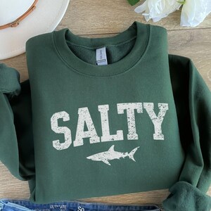 May include: A dark green sweatshirt with the word "SALTY" printed in white with a distressed look. A white shark silhouette is printed below the word.