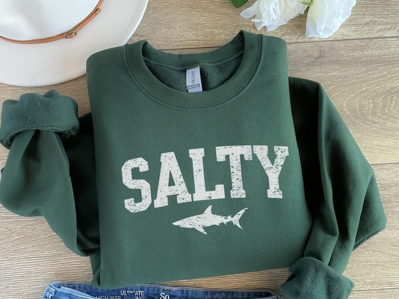 May include: A dark green sweatshirt with the word "SALTY" printed in white with a distressed look. A white shark silhouette is printed below the word.