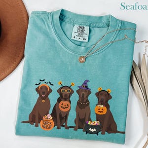 Pode incluir: Camiseta verde-água com um design de Halloween de quatro cães marrons fantasiados. Os cães usam chapéus de bruxa, tiaras de abóbora e óculos. Um cão segura um balde de doces "Trick or Treat".