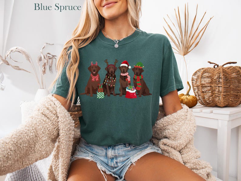 Op de afbeelding: Een tealkleurig T-shirt met bovenaan de tekst "Blue Spruce". Het shirt heeft een afbeelding van vier bruine Labrador Retrievers met feestelijke vakantieaccessoires. E&eacute;n hond draagt een rendiergeweige haarband, een ander heeft kerstverlichting en twee dragen kerstmutsen.