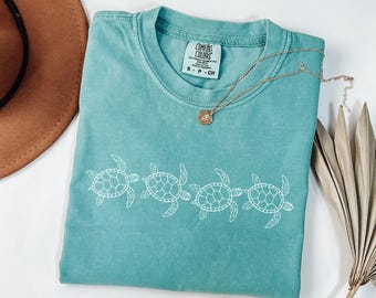 May include: A teal blue t-shirt with a white graphic of four sea turtles. The shirt has a tag that reads "COMFORT COLORS" and "S - P - CH".