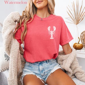 以下が含まれることがあります： 白いロブスターのグラフィックがプリントされたピンクのTシャツを着た女性。Tシャツはくたびれた感じ。デニムのショートパンツと白いふわふわのカーディガンも着ている。