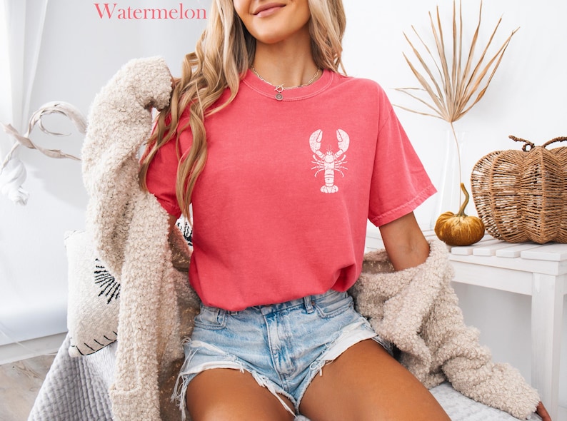 以下が含まれることがあります： 白いロブスターのグラフィックがプリントされたピンクのTシャツを着た女性。Tシャツはくたびれた感じ。デニムのショートパンツと白いふわふわのカーディガンも着ている。