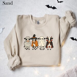 May include: Sand-colored sweatshirt featuring a Halloween-themed design. The graphic depicts four cartoon cows dressed as a mummy, witch, ghost, and vampire. Black bats and string lights decorate the background. A cozy and festive apparel item.