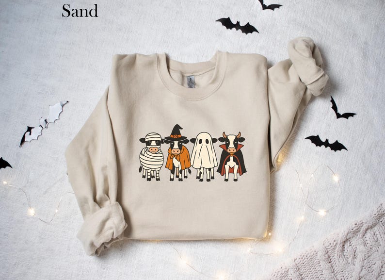May include: Sand-colored sweatshirt featuring a Halloween-themed design. The graphic depicts four cartoon cows dressed as a mummy, witch, ghost, and vampire. Black bats and string lights decorate the background. A cozy and festive apparel item.