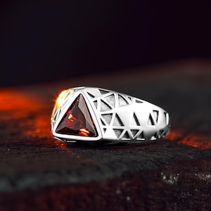 May include: A silver ring with a geometric design and a red gemstone in the center.