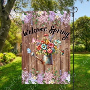 Puede incluir: Una bandera de jardín con fondo de madera y las palabras "Welcome Spring" en escritura negra. La bandera presenta una regadera rebosante de flores coloridas y mariposas. Flores moradas y vegetación enmarcan la parte superior e inferior de la bandera.