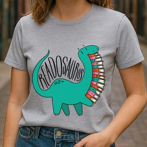 May include: Ash gray t-shirt featuring a teal dinosaur graphic with the word "READOSAURUS" and a stack of colorful books. The shirt is short-sleeved and made of a soft material. The word "Ash" is printed in white in the bottom left corner.
