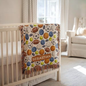 Op de afbeelding: Een beige babydeken met een patroon van verschillende sportballen, waaronder American footballs, voetballen, basketballen en tennisballen. De deken heeft een bruine patch met de naam "SEBASTIAN". De deken ligt over een wit babybed.
