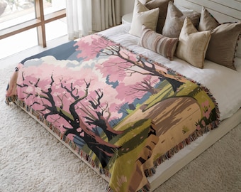 May include: A woven throw blanket with a scenic illustration of cherry blossom trees in full bloom. The design includes pink blossoms, dark tree trunks, and a landscape with green and brown tones. The blanket has a fringed edge.