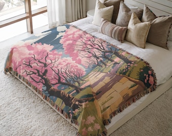 May include: A woven throw blanket with a scenic illustration of a cherry blossom landscape. The design includes pink flowering trees, a path, and a blue sky. The blanket has a fringed edge and is displayed on a bed with pillows.