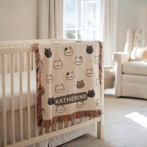 May include: A beige baby blanket with a repeating pattern of cat faces in various colors. The blanket has a fringed edge and the name "KATHERINE" printed on it. The blanket is draped over a white crib.