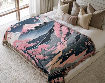 May include: A woven throw blanket with a Japanese landscape design, depicting a mountain, cherry blossom trees, and a river. The colour scheme includes pink, blue, grey, and black. The blanket has a fringed edge and is shown on a bed.