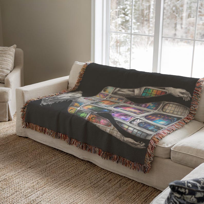 Puede incluir: Una manta negra con una ilustraci&oacute;n detallada de una figura con patrones geom&eacute;tricos coloridos. La manta tiene un borde con flecos y est&aacute; colocada sobre un sof&aacute; blanco. La obra de arte presenta una mezcla de colores y formas.