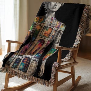 Puede incluir: Una manta negra con una ilustraci&oacute;n detallada de una figura con ventanas que representan paisajes coloridos. La manta est&aacute; sobre una mecedora de madera y tiene un borde con flecos. La manta est&aacute; hecha de un material suave.