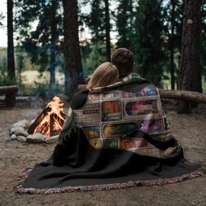 Puede incluir: Una manta negra con un dise&ntilde;o estampado colorido sobre dos personas sentadas cerca de una fogata. La manta tiene un borde con flecos de varios colores. El dise&ntilde;o de la manta presenta m&uacute;ltiples paneles rectangulares con im&aacute;genes coloridas.