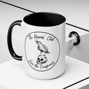 May include: White ceramic mug with a black interior, handle, and rim. The mug features a circular design with a raven perched on a skull, and the text "The Ravens' Club" and "Join the Conspiracy" in a gothic font.