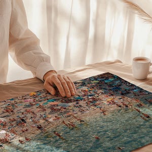 May include: A close-up of a colorful jigsaw puzzle depicting a beach scene with people swimming and sunbathing in the ocean. The puzzle is laid out on a beige surface with a person's hand in the background.