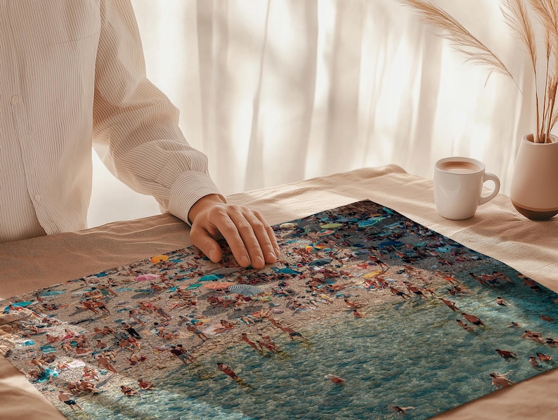 May include: A close-up of a colorful jigsaw puzzle depicting a beach scene with people swimming and sunbathing in the ocean. The puzzle is laid out on a beige surface with a person's hand in the background.