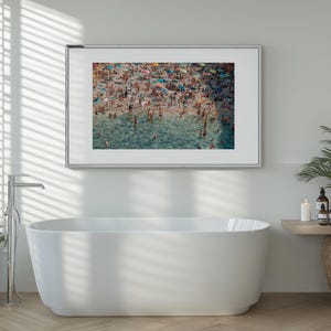 May include: A framed photograph of a crowded beach with people swimming and sunbathing in the turquoise water. The photo is hanging on a white wall above a white bathtub.