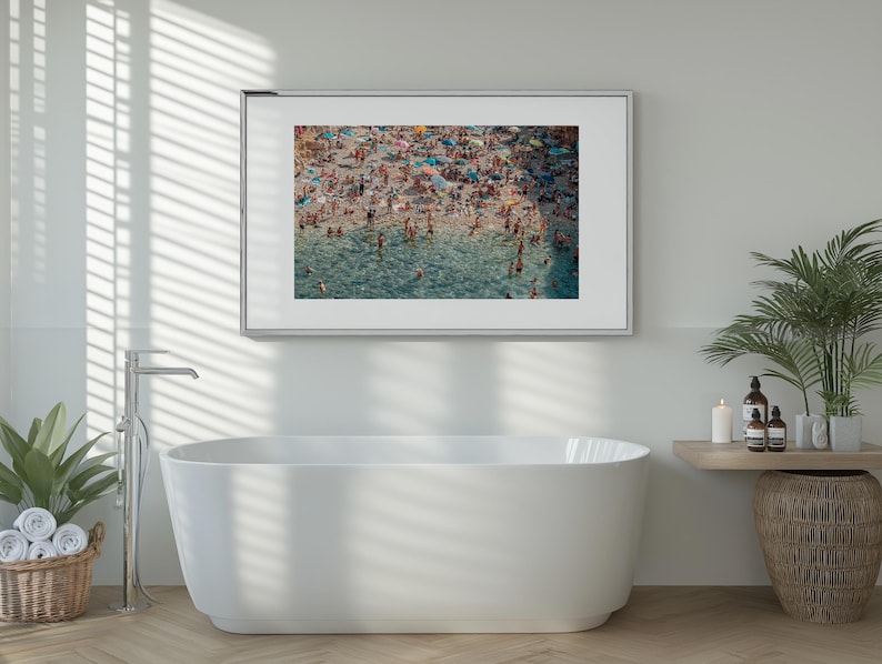 May include: A framed photograph of a crowded beach with people swimming and sunbathing in the turquoise water. The photo is hanging on a white wall above a white bathtub.
