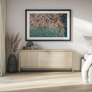May include: A framed photograph of a crowded beach scene with people swimming and sunbathing in the turquoise water. The photo is hanging on a white wall above a light wood dresser.